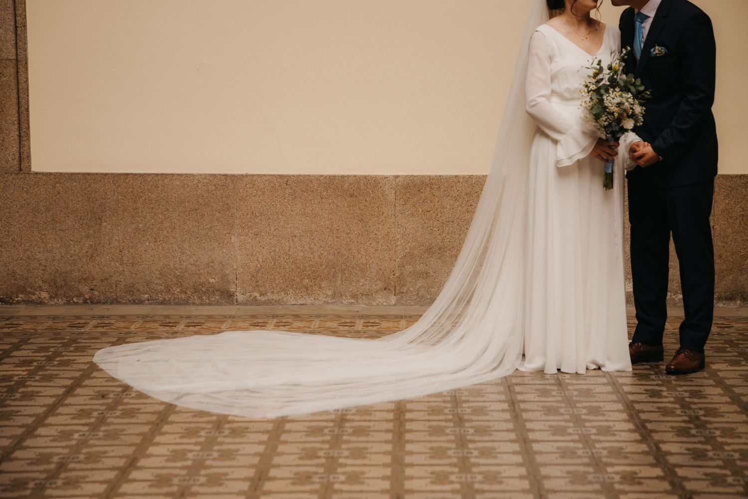 A long flowing wedding dress train against a cobblestone path with the couple standing together holding a bouquet.