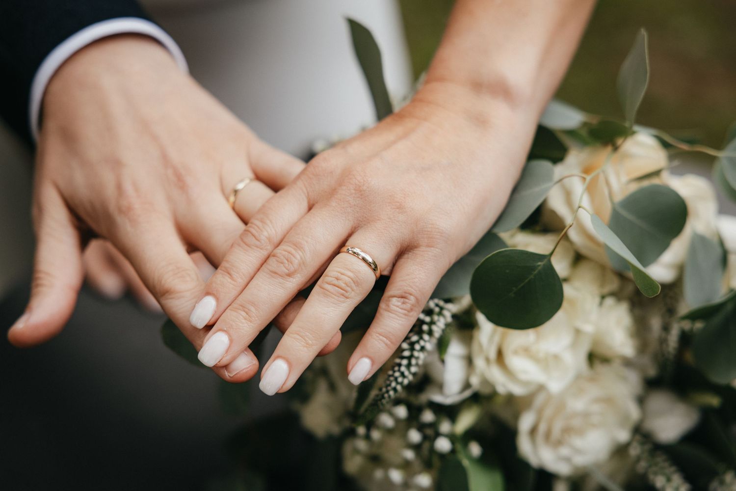 Close-up of wedding rings and hands with white roses and eucalyptus bouquet in the background.