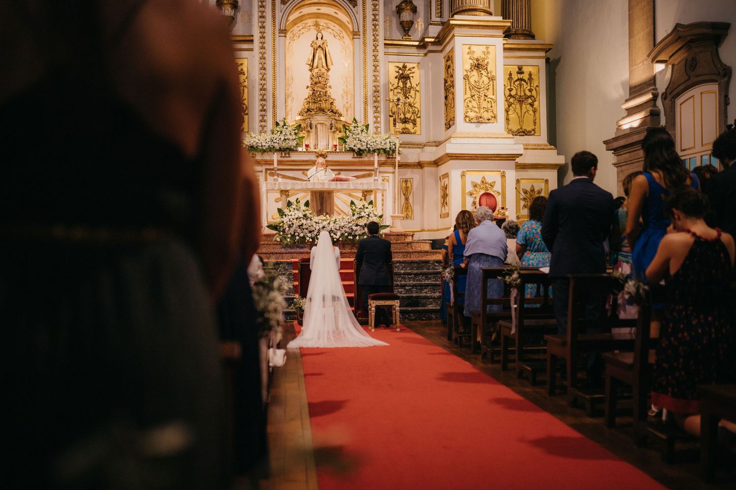 Wedding ceremony taking place in an ornate Catholic church with a red carpet aisle and gold altar decorations.
