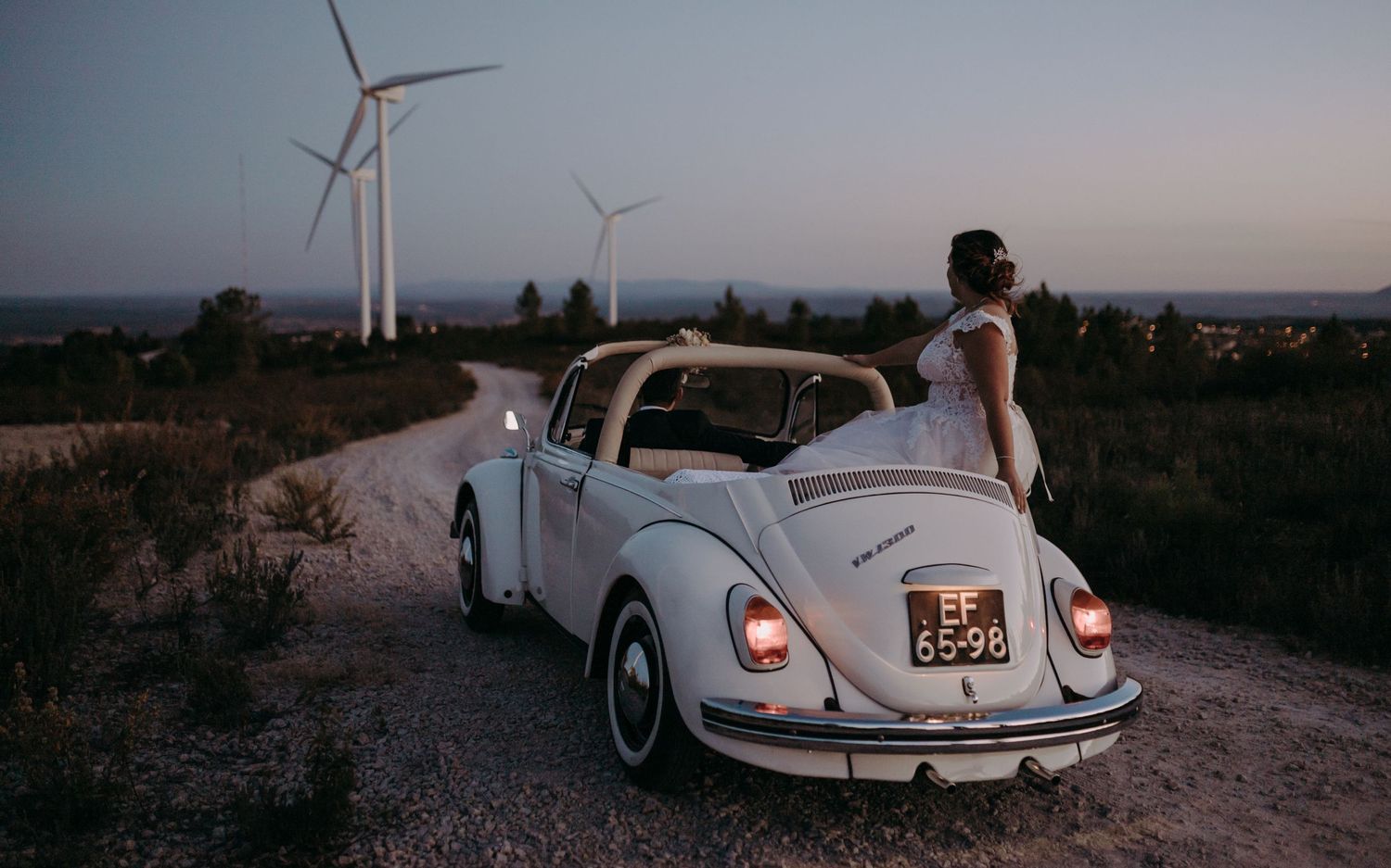 Vintage white Volkswagen Beetle parked on rural road with wind turbines in background at sunset.