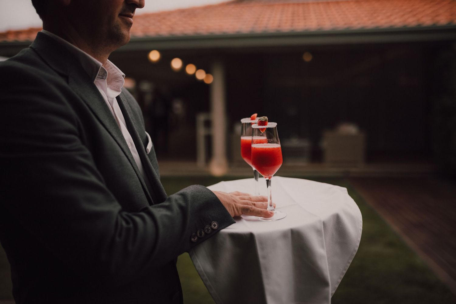 Wedding guest holds red cocktail on white linen against evening setting.