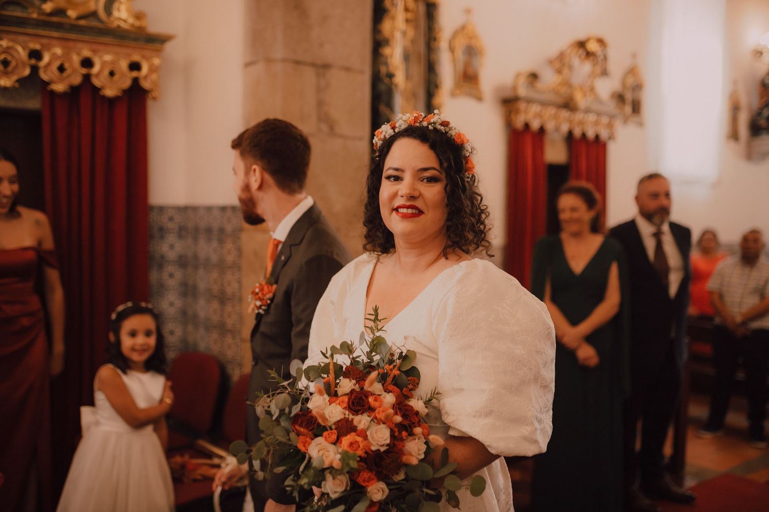 A joyful bride wearing a white dress with puff sleeves holds a vibrant orange and pink bouquet in an ornate church setting.