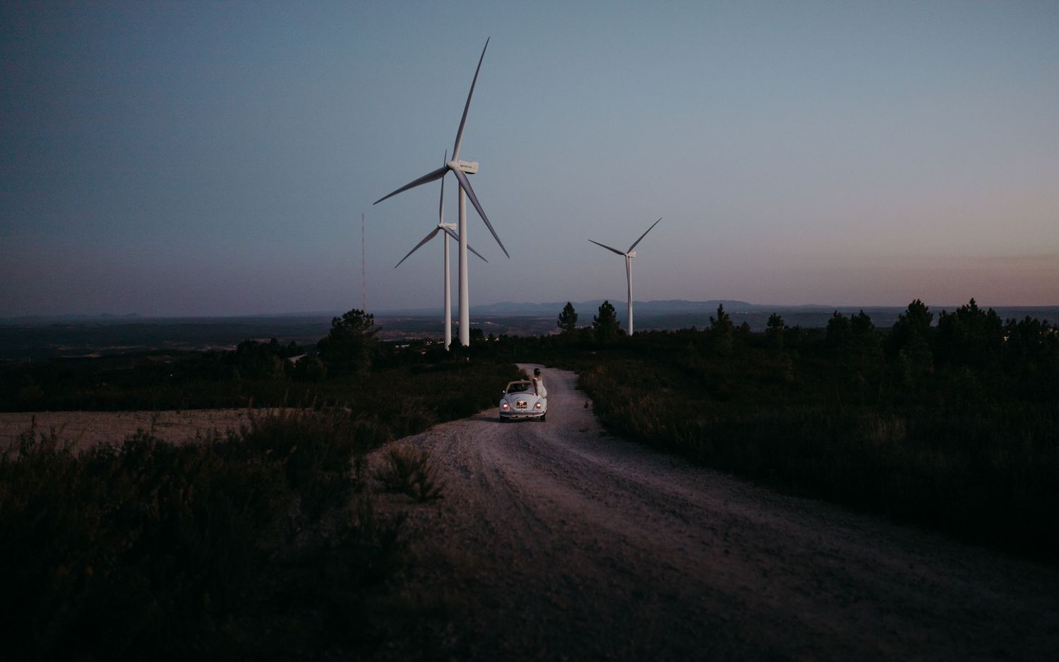 Classic white car driving on dirt road with wind turbines silhouetted against dusky sky.