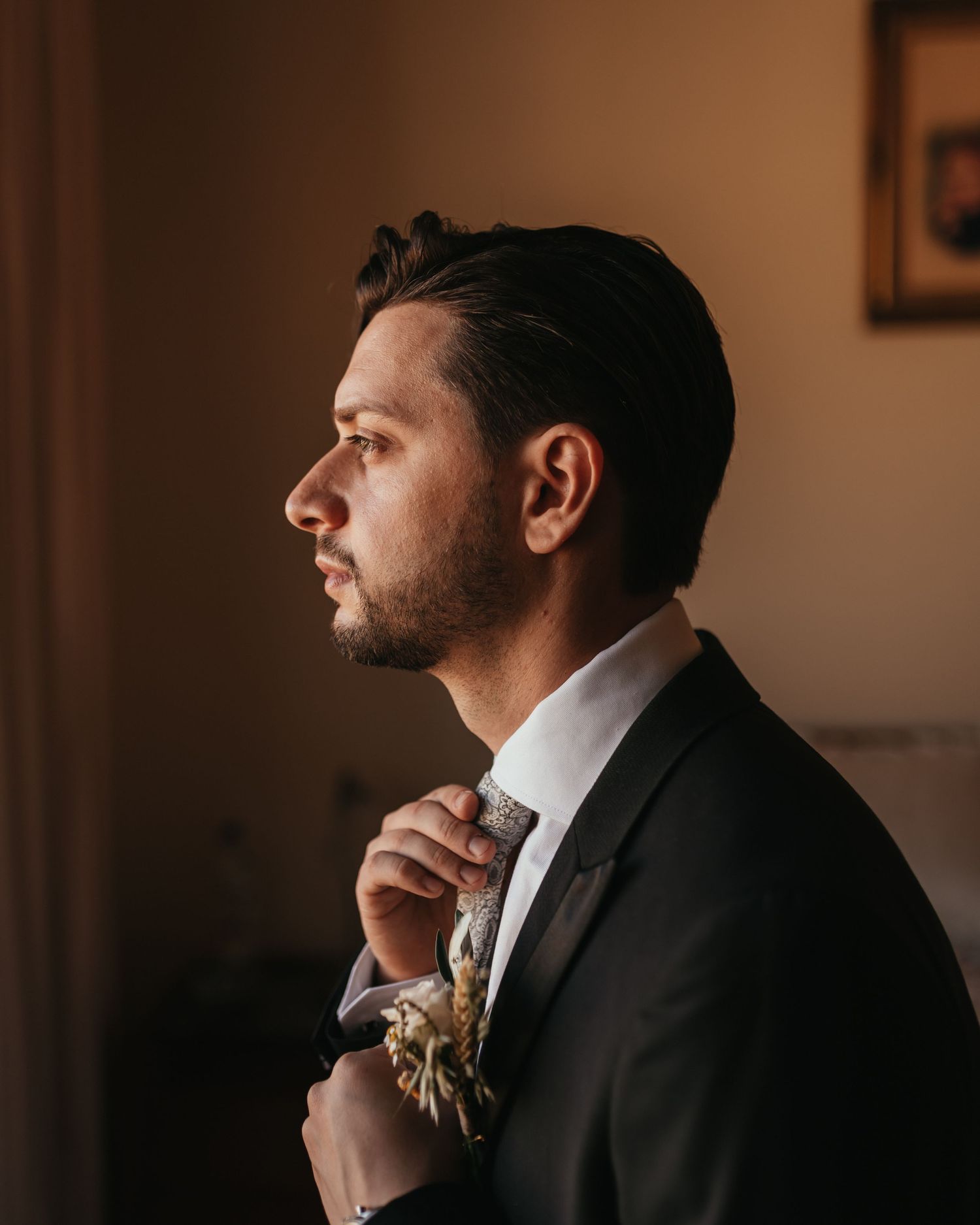 Side profile of groom in formal attire adjusting his tie in warm indoor lighting.