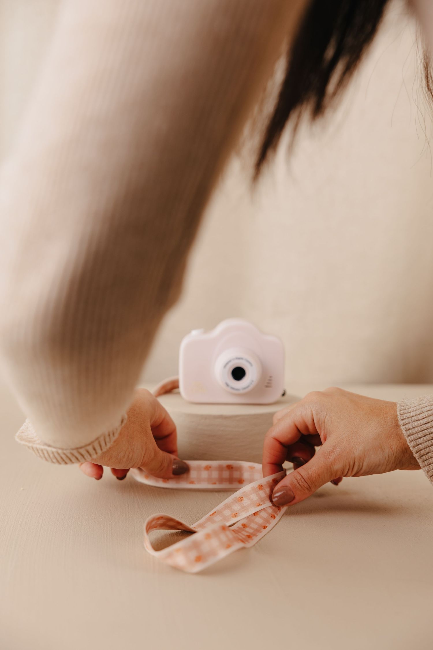 Hands holding a pink instant camera and photo strip on a beige surface with soft lighting.