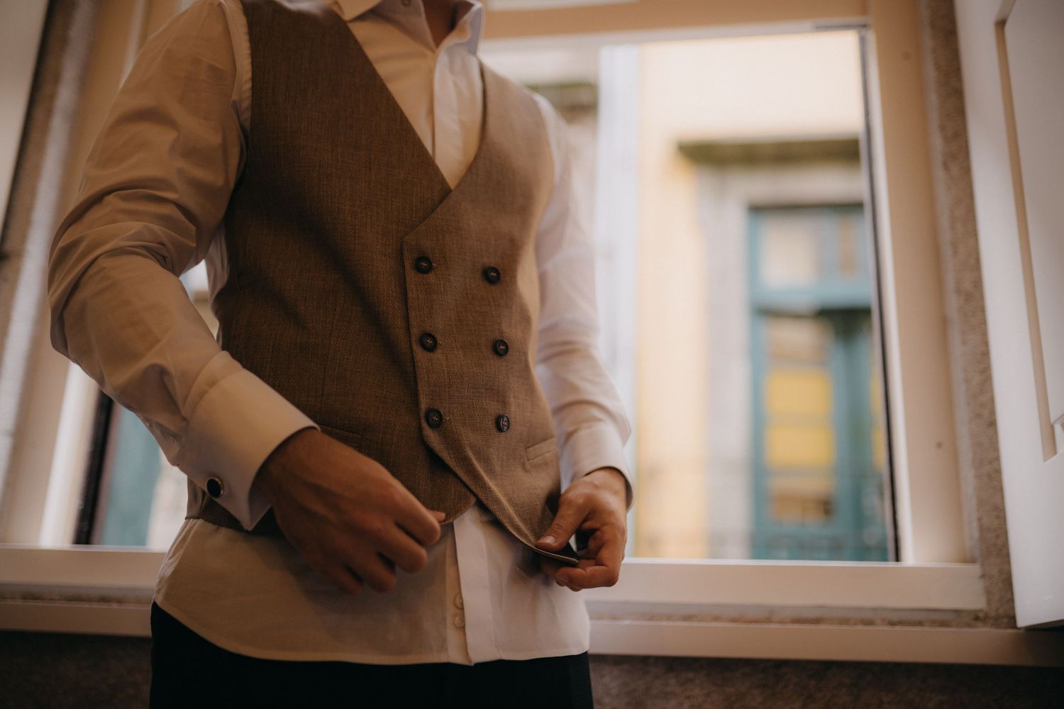Person wearing a beige waistcoat adjusts formal attire by a window.