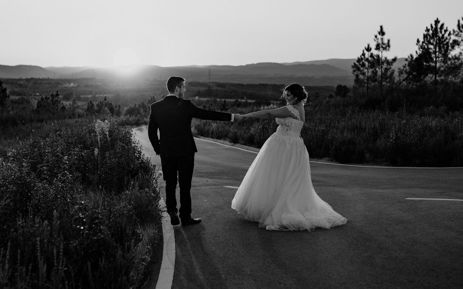 Bride and groom standing on a rural road at sunset with mountain views in black and white.