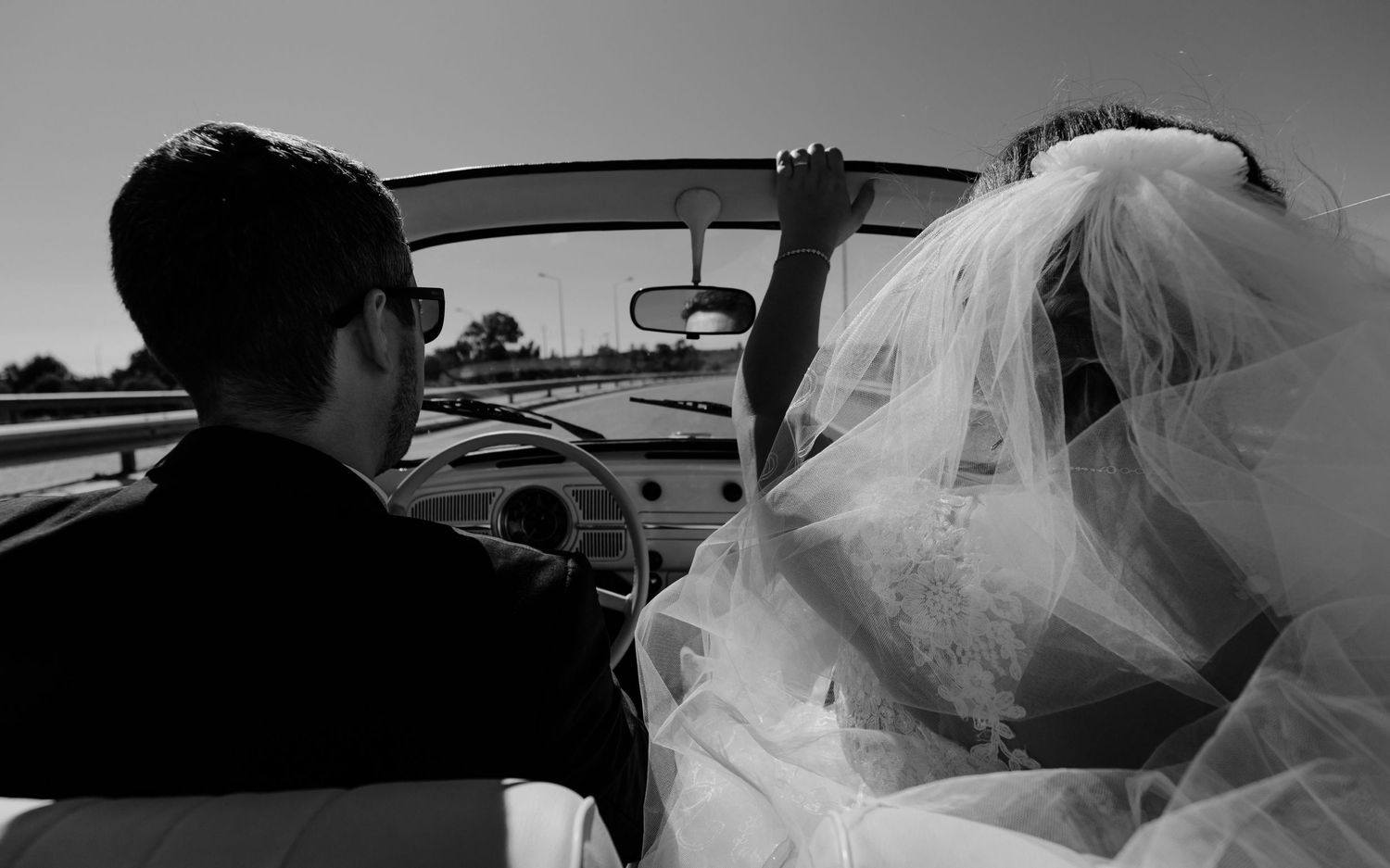 A black and white photo showing newlyweds in a vintage convertible car with flowing veil.
