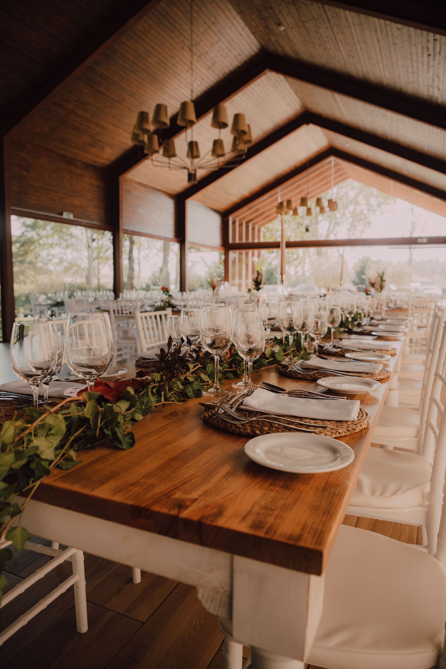 Elegant wedding reception table setting with rustic wooden tables and white chairs under a vaulted ceiling.