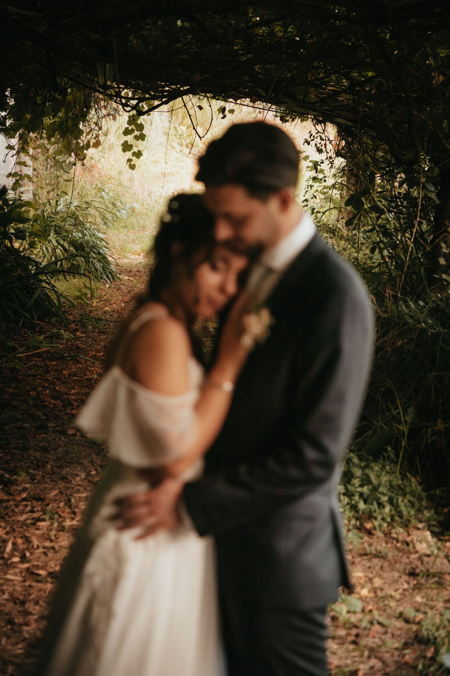 A romantic wedding couple embraces under a dreamy forest archway with soft natural lighting.