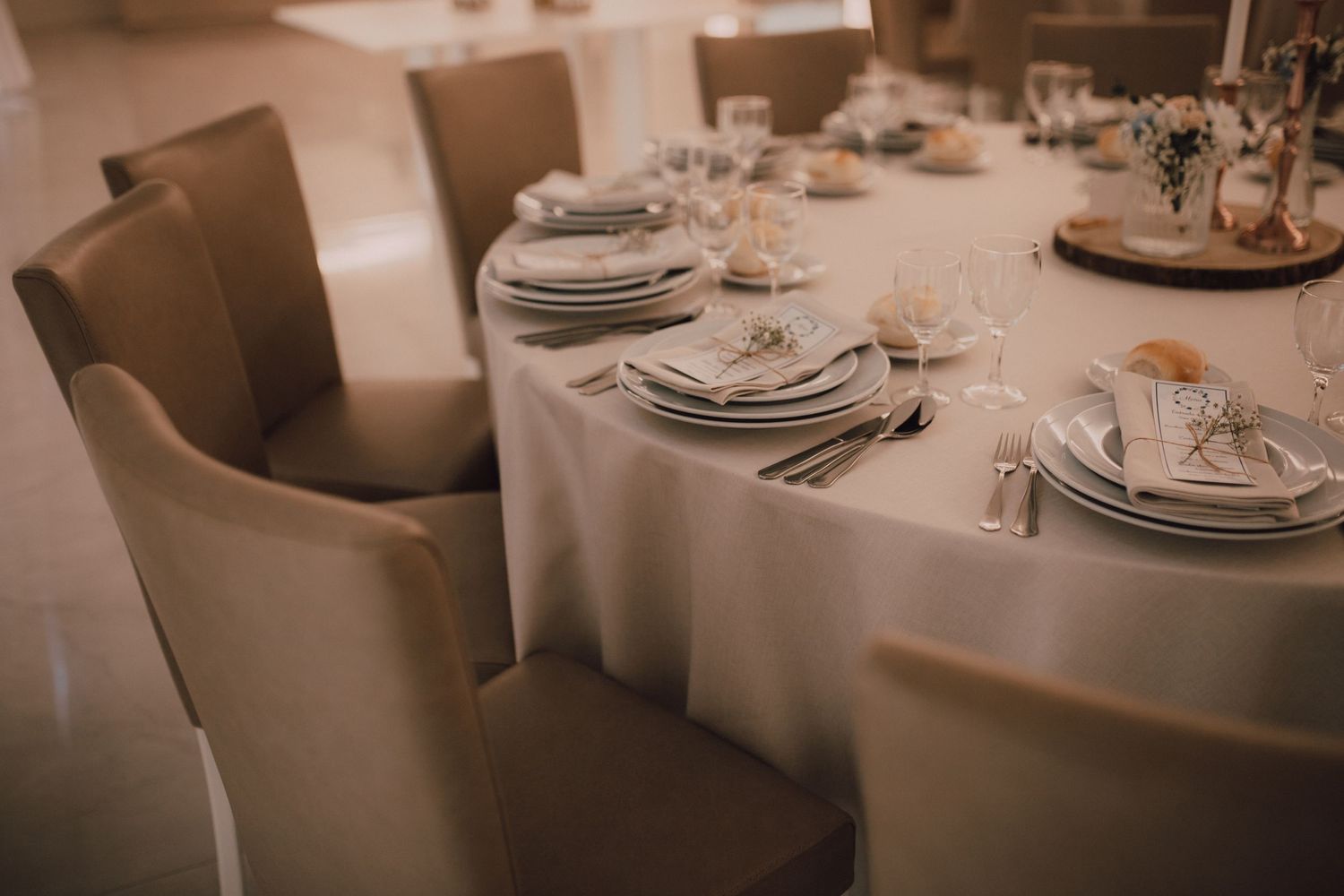 Formal dining table setup with white tablecloth and comfortable beige chairs in a reception hall.