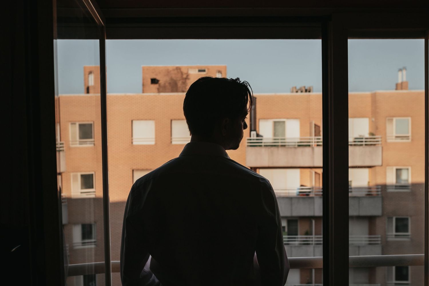 Silhouette by window overlooking urban apartment buildings at sunset.