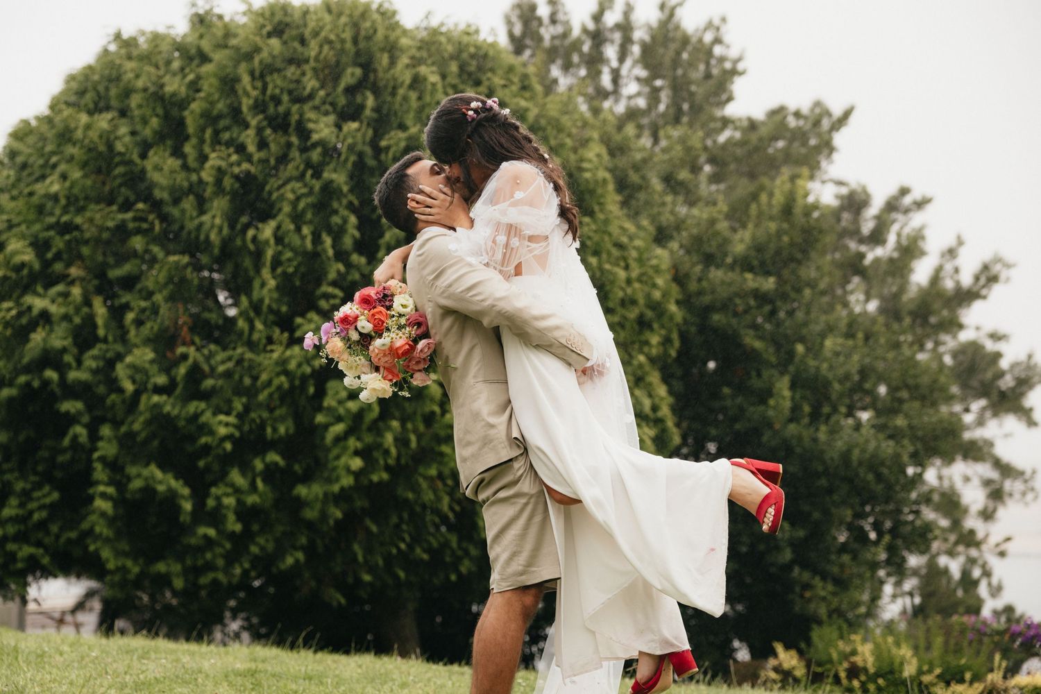 Bride in flowing white dress and red shoes being lifted during outdoor wedding celebration.