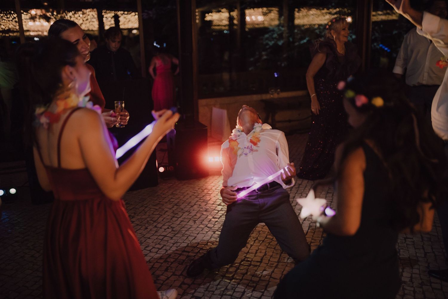 Wedding guests dance with glow sticks under dim lighting at an evening reception celebration.