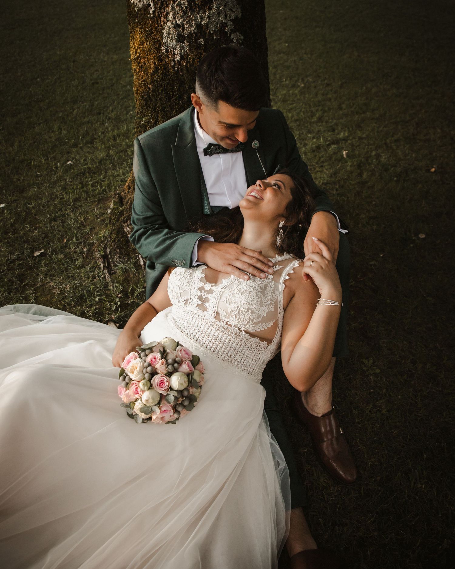 A romantic wedding photo shows a couple in formal attire with a pink bouquet resting against a tree trunk.