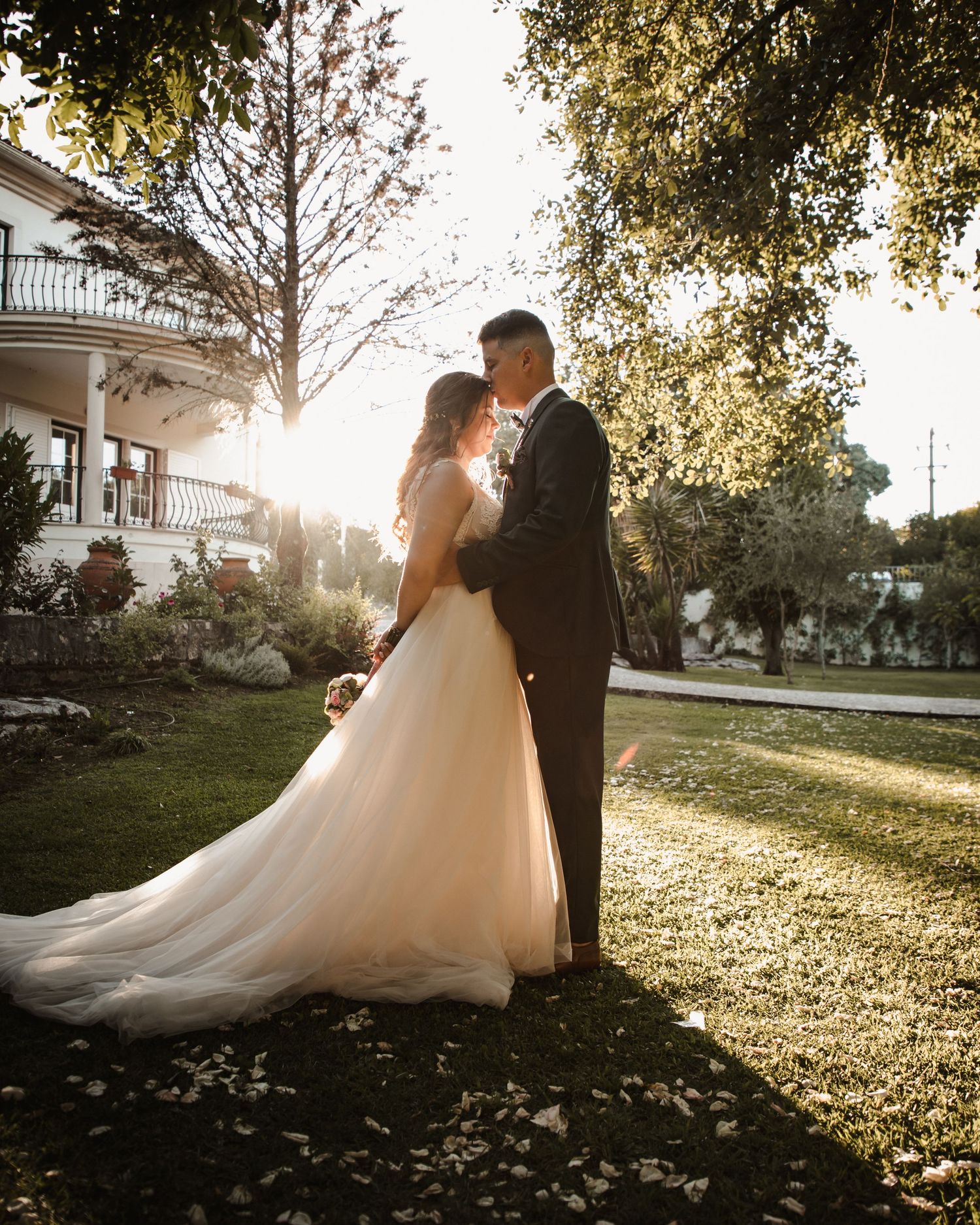 A wedding couple shares an intimate moment in golden sunlight on a manicured lawn.