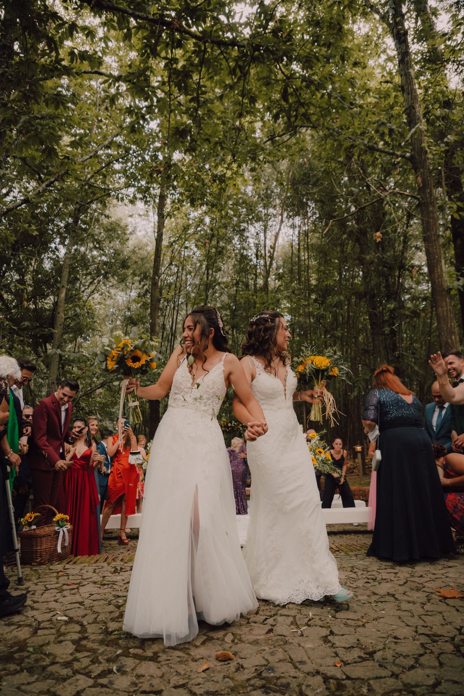 Two brides in white dresses celebrate with guests at an outdoor woodland wedding ceremony.