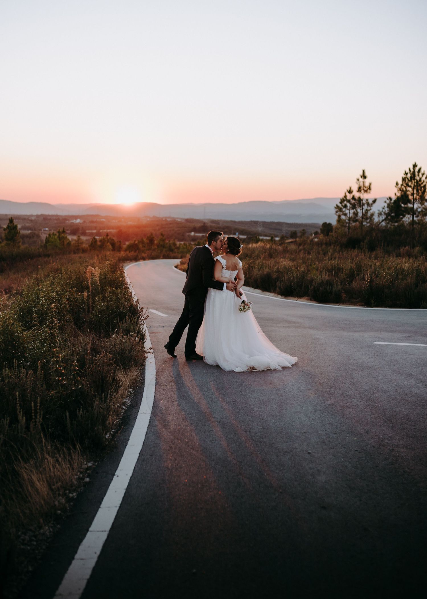 A wedding couple stands on a winding mountain road at sunset creating a romantic silhouette against the scenic landscape.