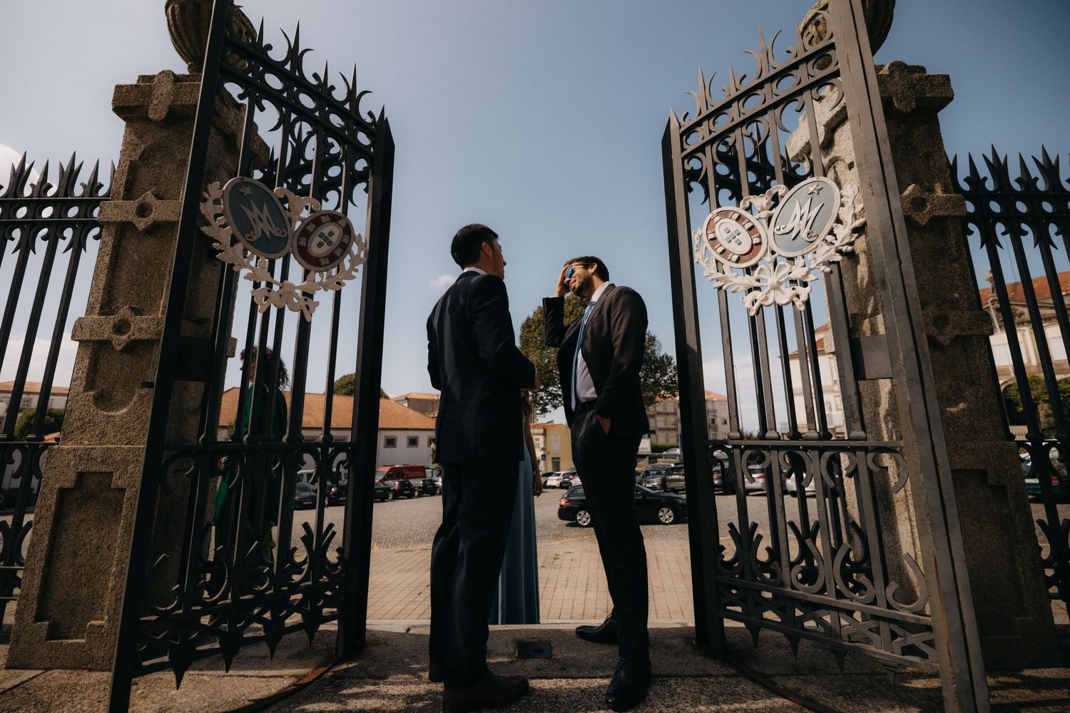 Two figures stand silhouetted between ornate iron gates during a moody outdoor scene.