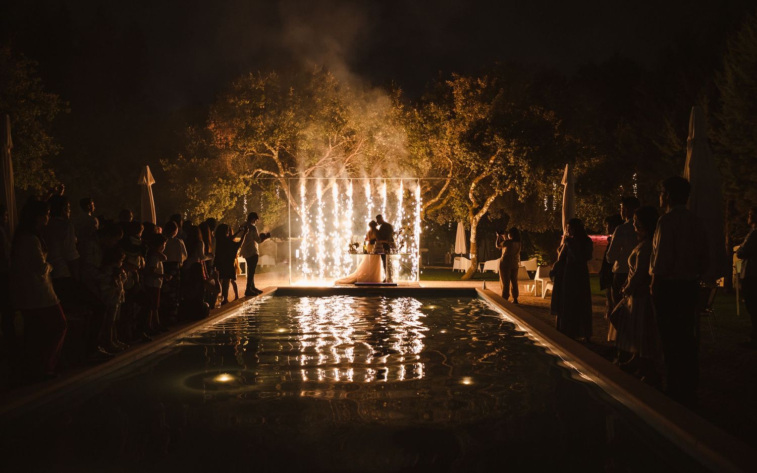 Illuminated wedding ceremony setup with glowing archway reflected in pool at night.