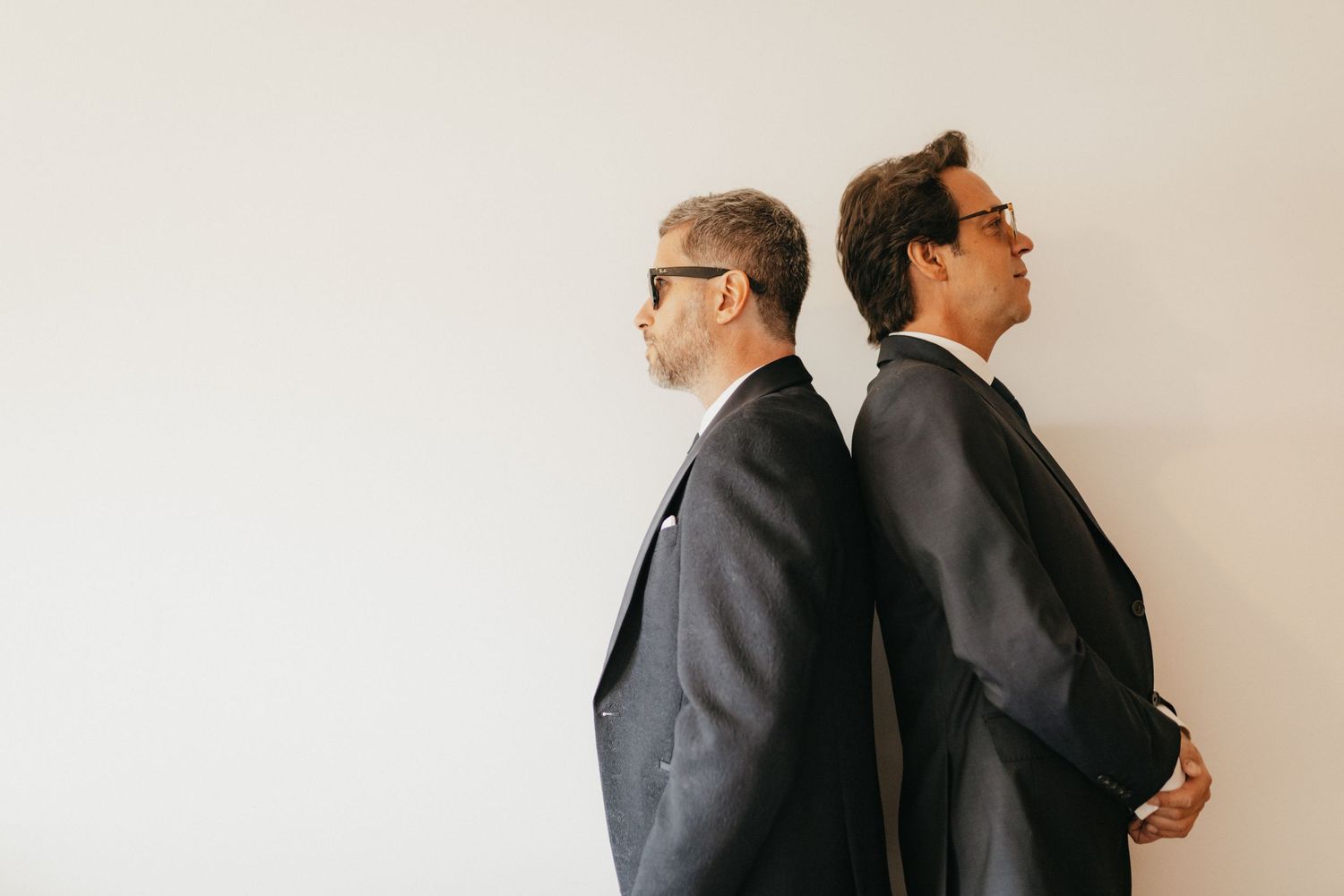 Two people in dark suits standing back to back against white wall in formal portraits.