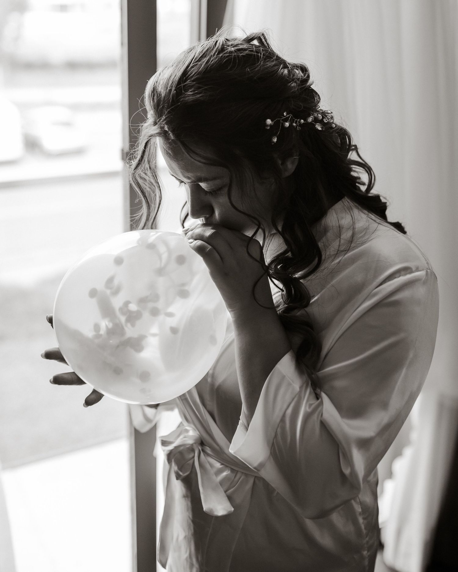 Wedding preparation moment as a bride in white robe blows up a balloon by the window.