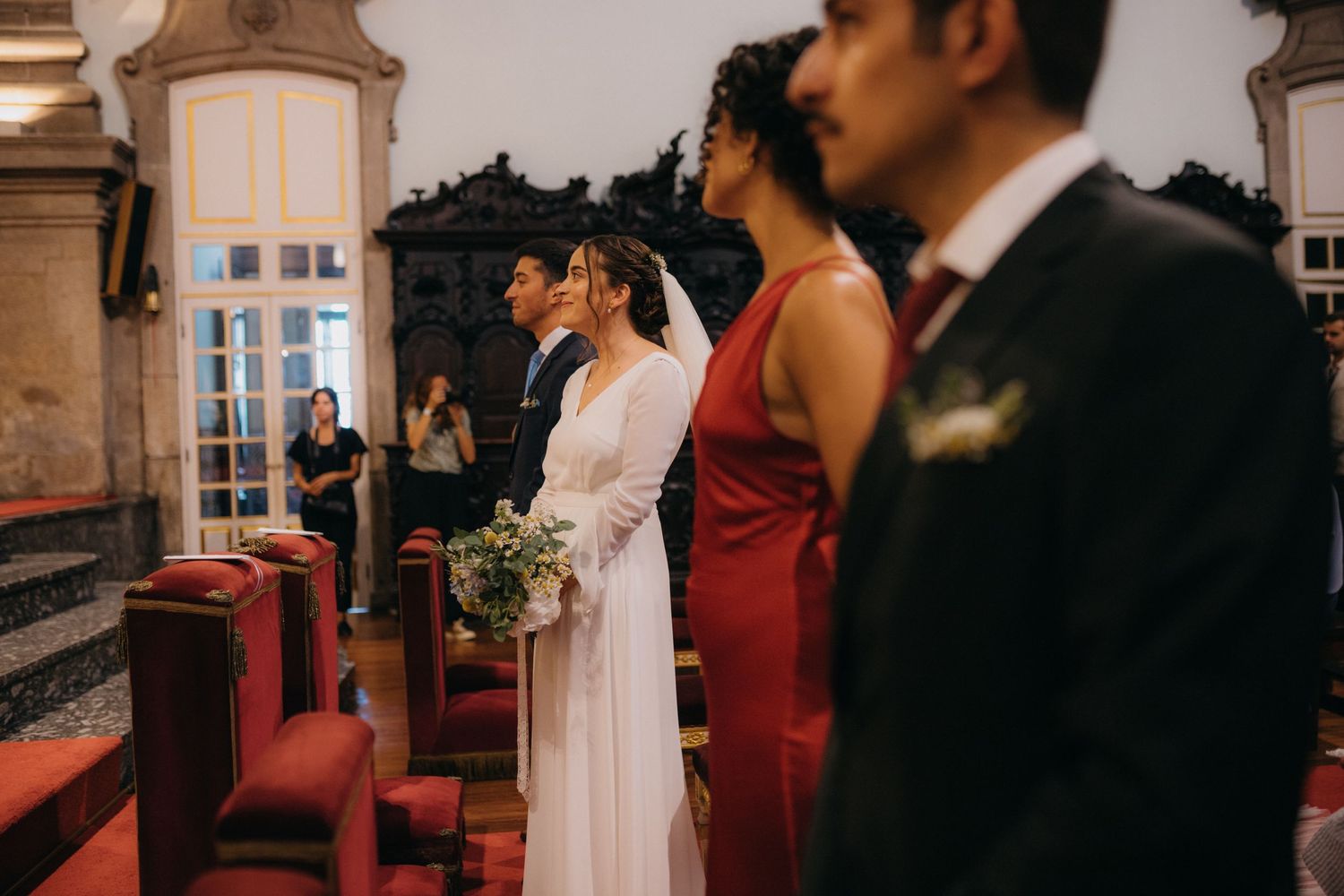 Wedding ceremony attendees stand in an ornate church interior featuring red carpet and traditional decor.