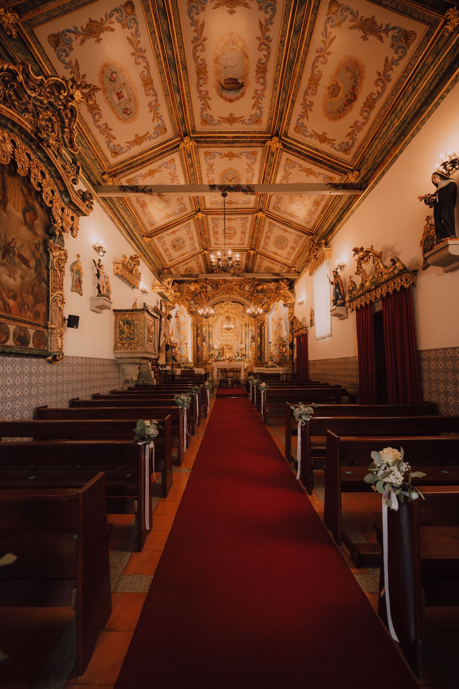Interior of a historic church with ornate ceiling frescoes, wooden pews, and red carpet aisle decorations.