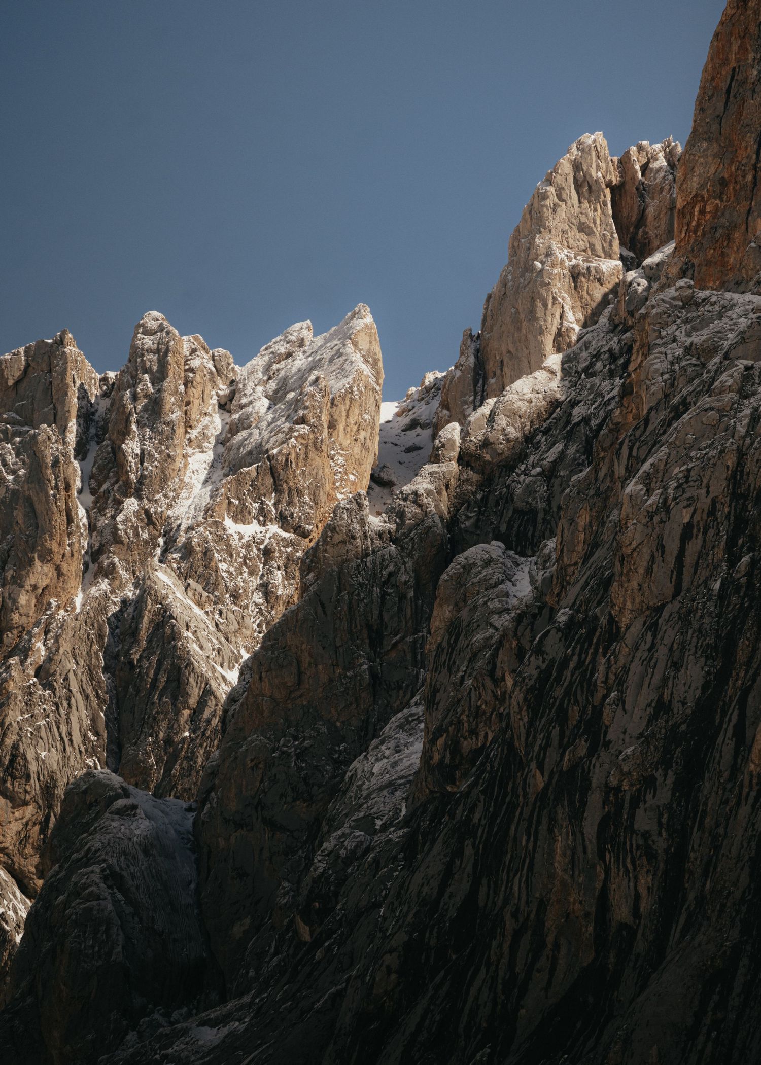 Sunlit rocky mountain peaks with steep granite cliffs and snow patches against a deep blue sky.
