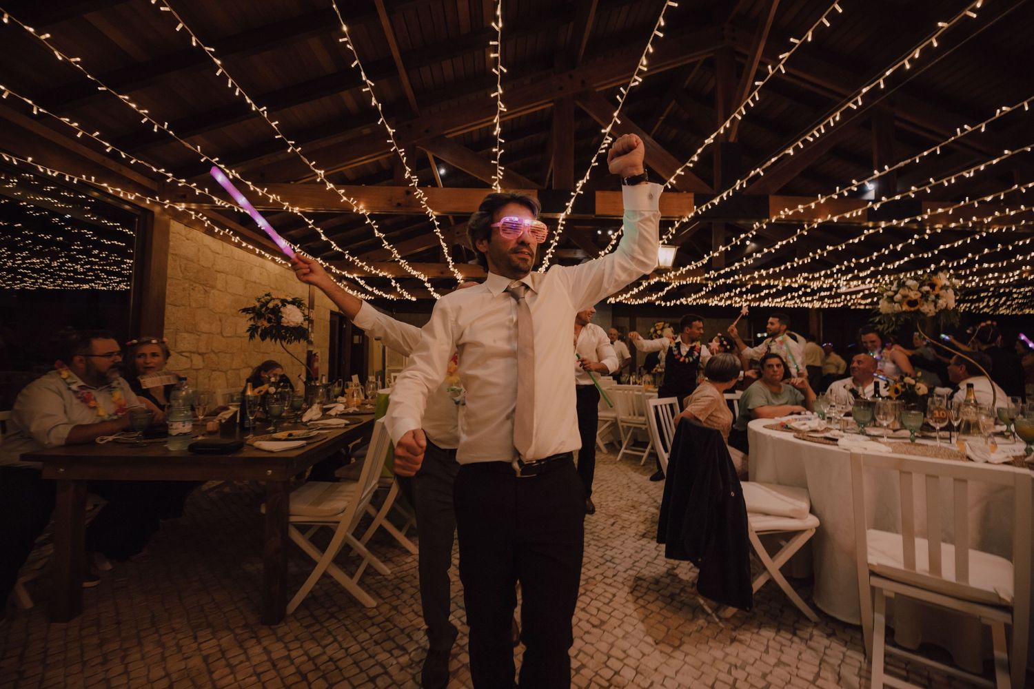 Guest raises arms in celebration under twinkling string lights at wedding reception.