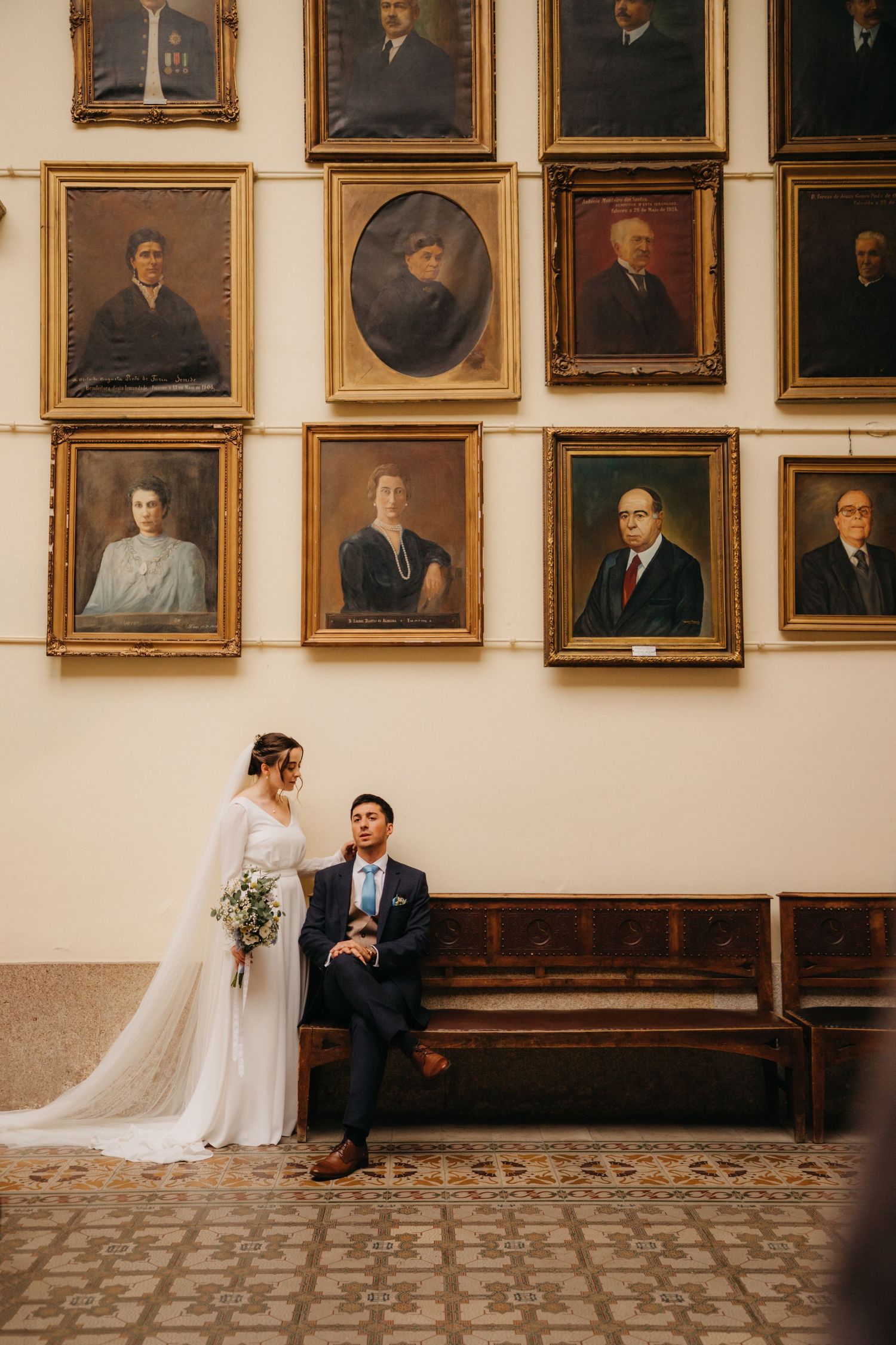 A couple sits on a wooden bench beneath a wall of historic portrait paintings.