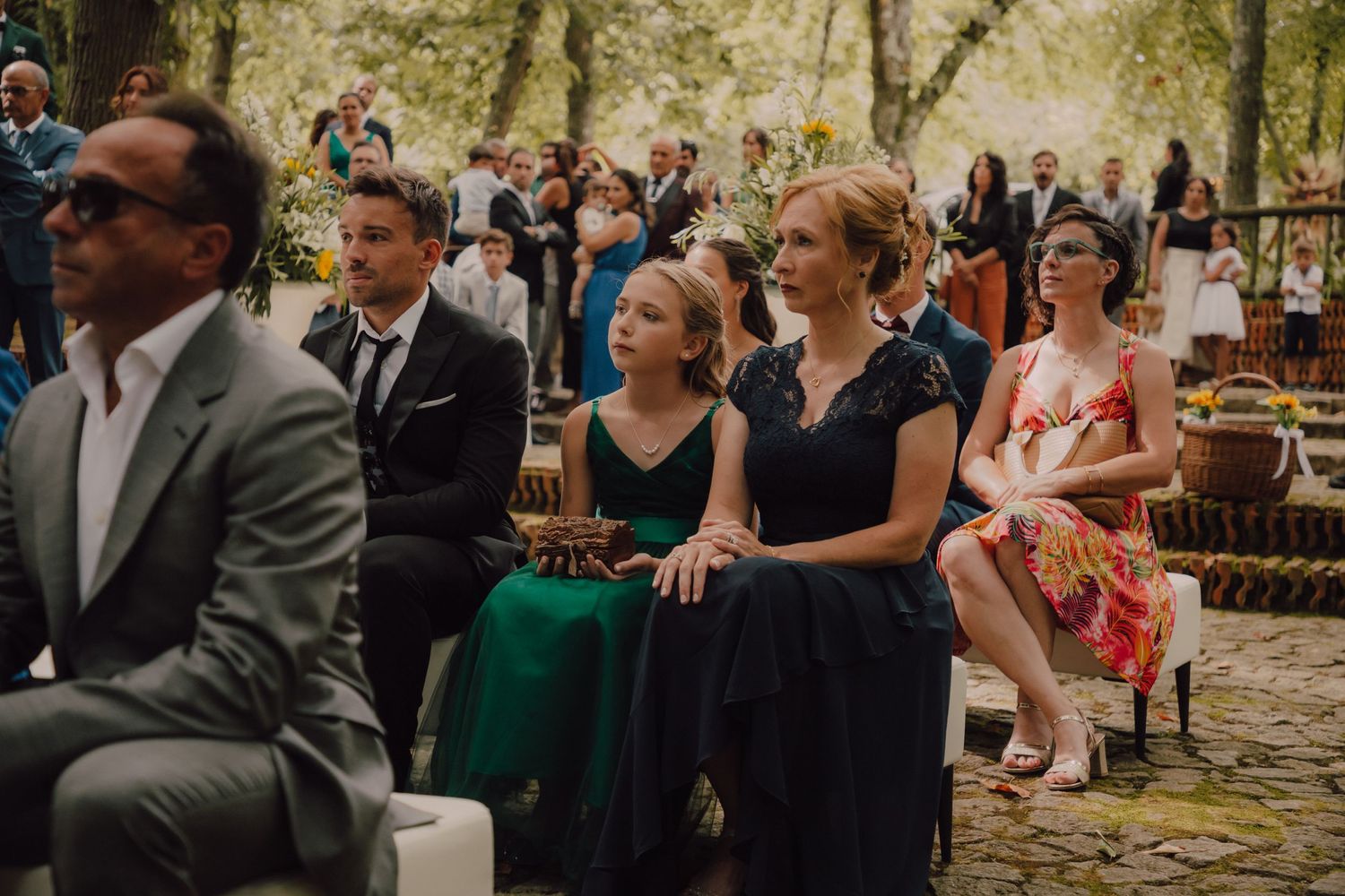 Wedding guests in formal attire seated outdoors at a ceremony with sunlit trees in the background.
