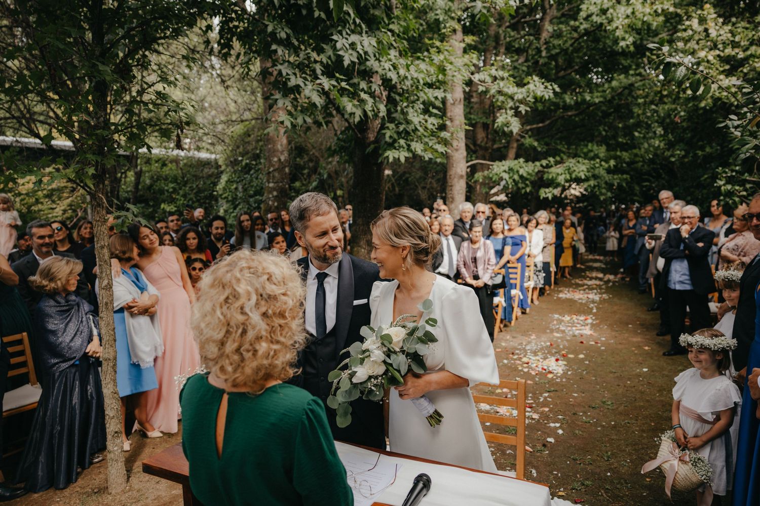 Outdoor wedding ceremony in a wooded area with guests gathered around chairs on a tree-lined path.