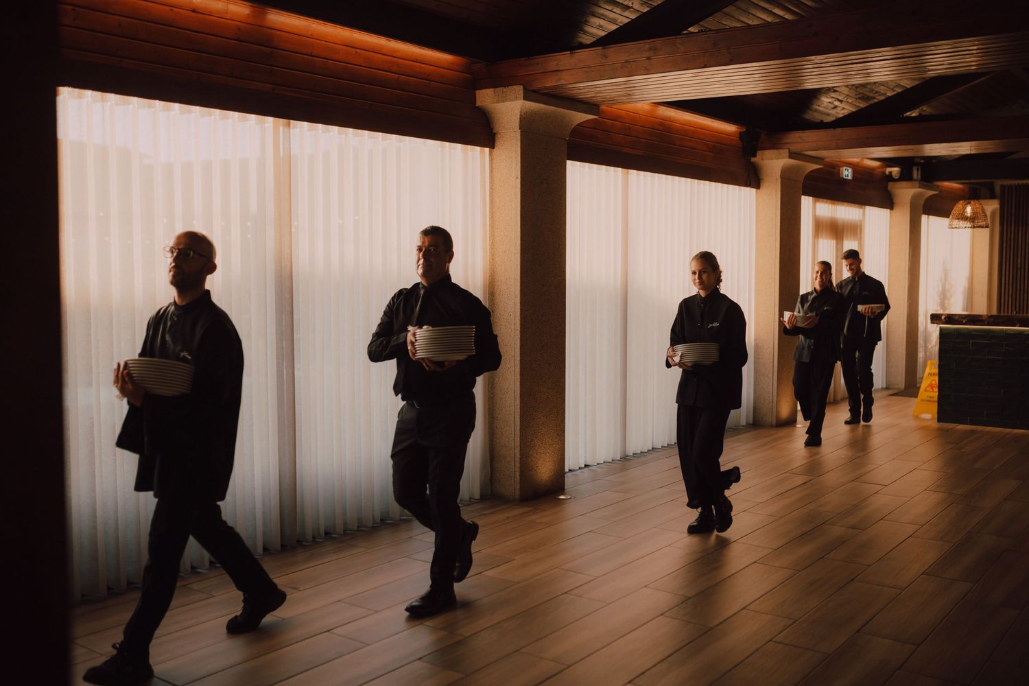 Service staff in black attire walk through a modern hallway with wooden floors.