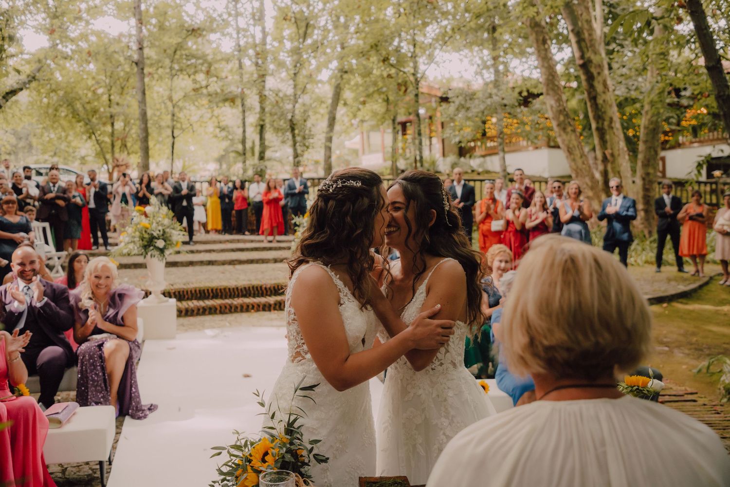Outdoor wedding ceremony with guests seated on white chairs among trees and natural decor.