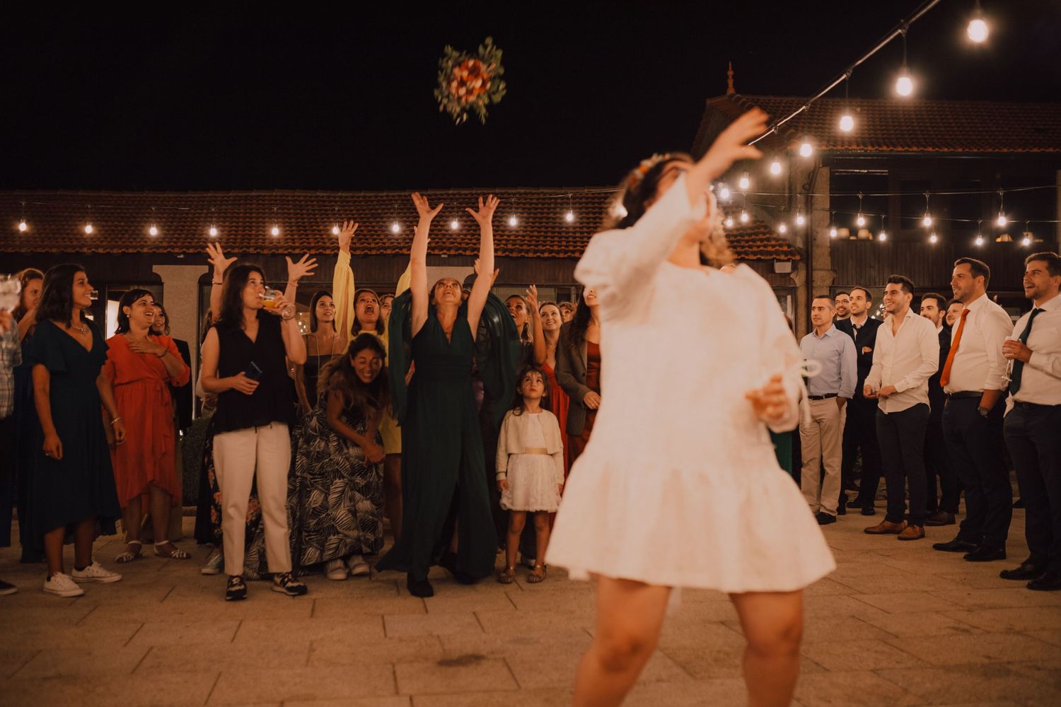 A bride tosses her bouquet during a nighttime wedding reception with string lights and excited guests.
