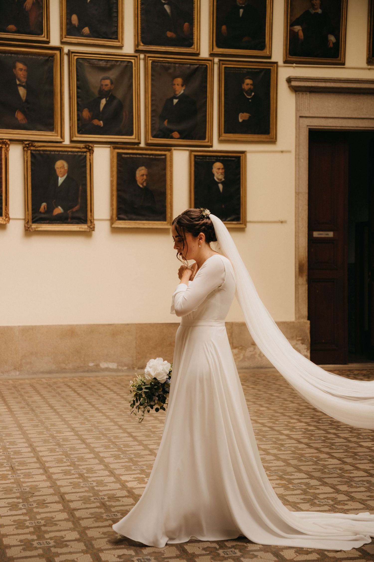 An elegant wedding dress with flowing veil photographed against a wall of historic portraits.