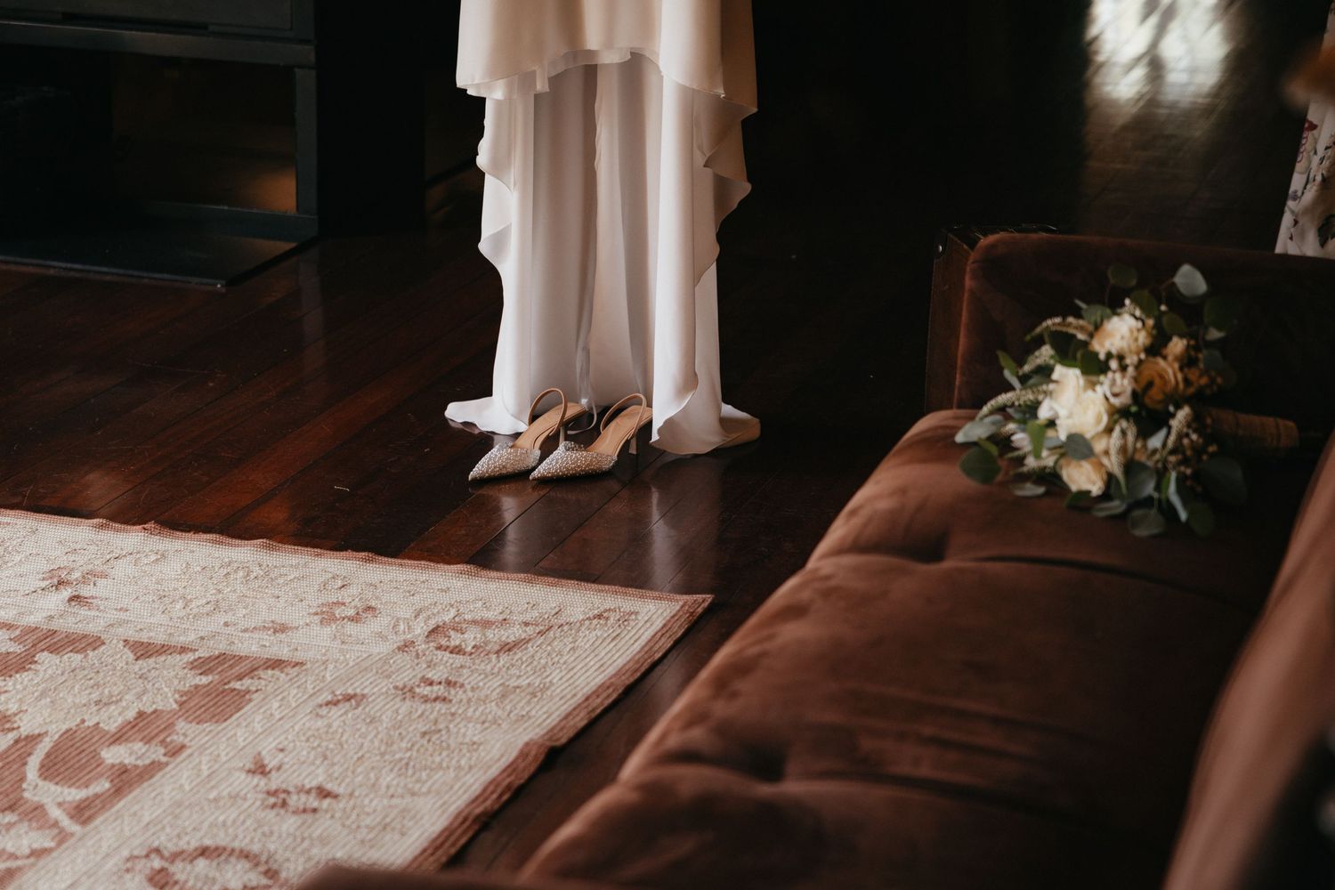Wedding dress hanging with bridal shoes and bouquet on brown sofa in natural light.