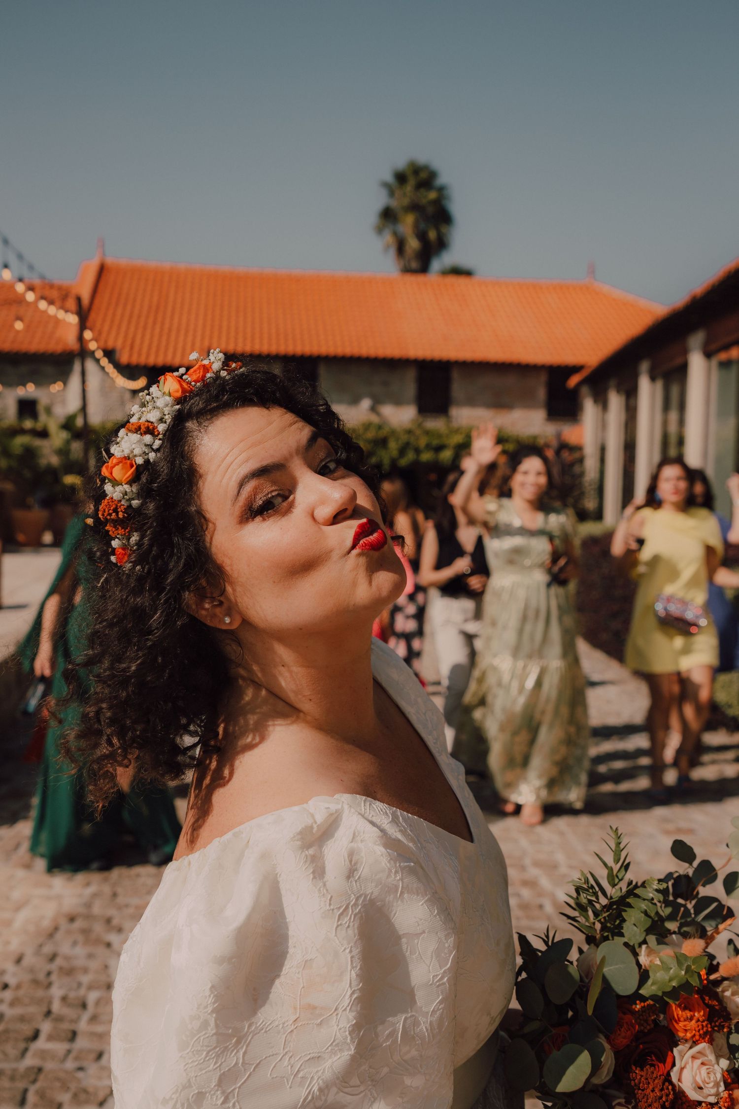 A person in a white off-shoulder dress with floral crown poses outdoors against terracotta roofs and palm trees.