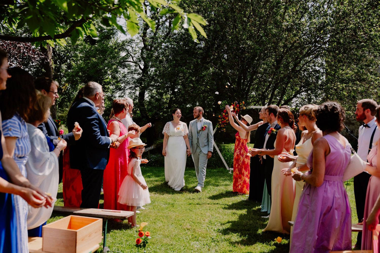 Outdoor wedding ceremony with guests forming an aisle, tossing petals at the couple on a sunny green lawn.