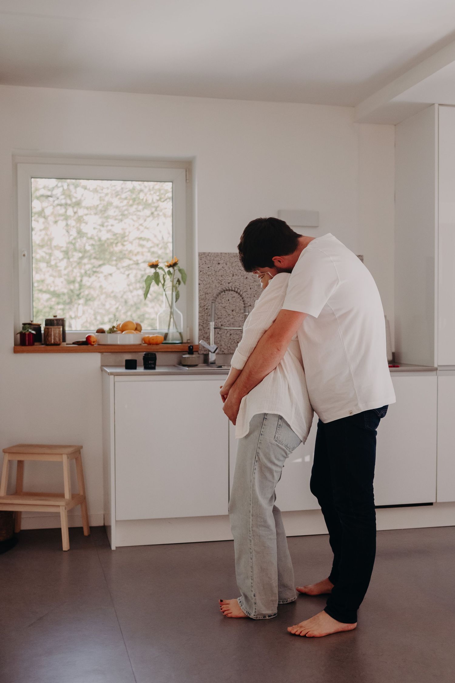 Two people standing barefoot in a minimalist white kitchen with a window, one helping support the other.