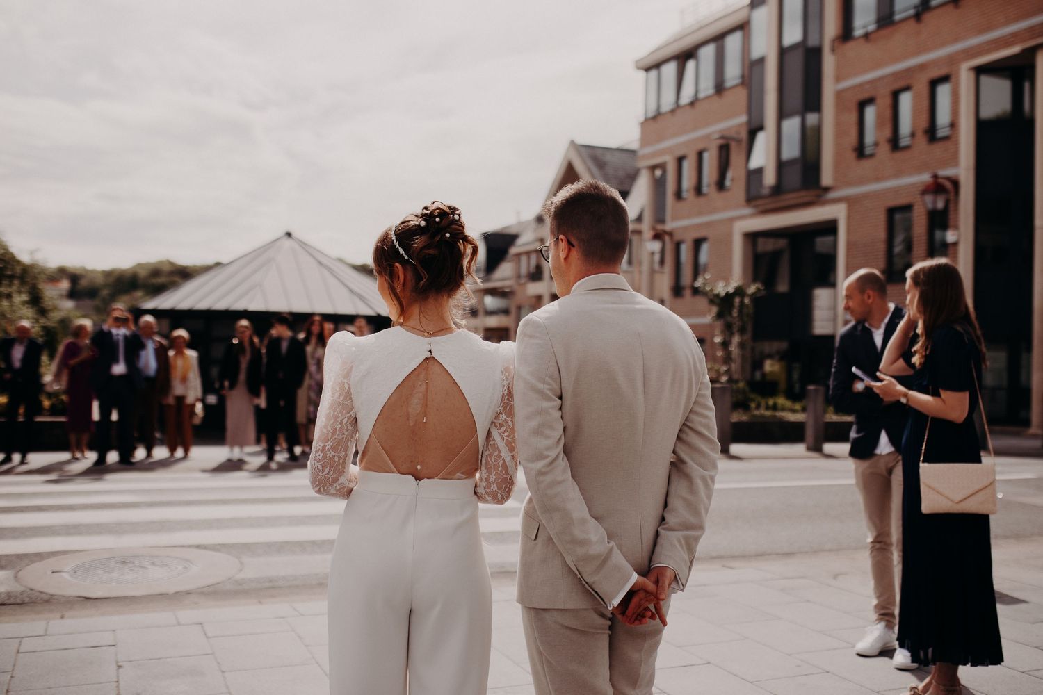 A couple in white wedding attire stands together at an outdoor venue with guests and tents in the background.