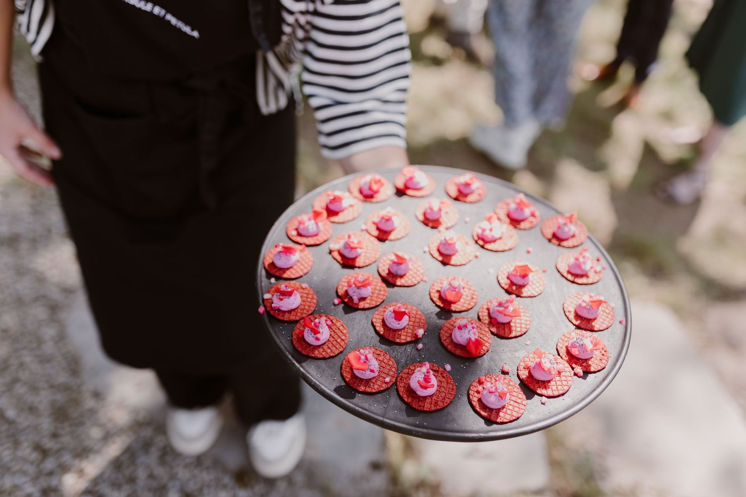 A silver platter displays multiple pink frosted mini cupcakes decorated with white sprinkles being held at an outdoor event.