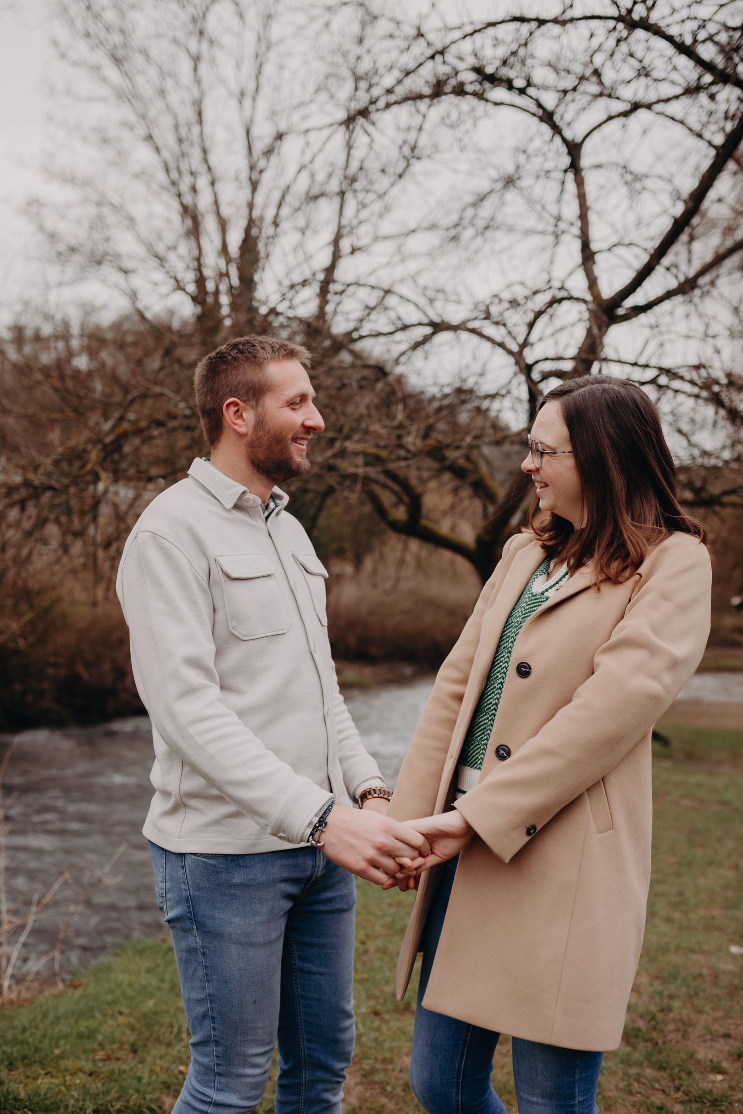 A couple holding hands and smiling at each other in a park during autumn with bare trees and a stream in the background.