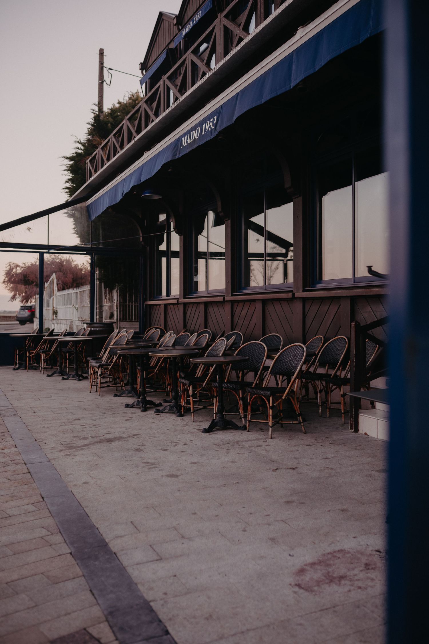 Empty bistro chairs line the sidewalk outside a French cafe with blue awnings at dusk.