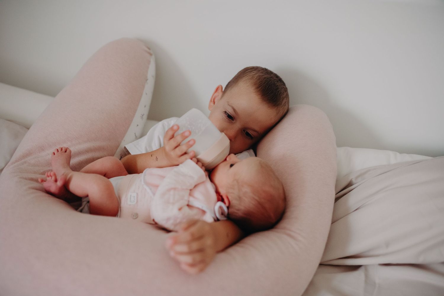 A baby in a white onesie lies on a curved nursing pillow while drinking from a bottle.