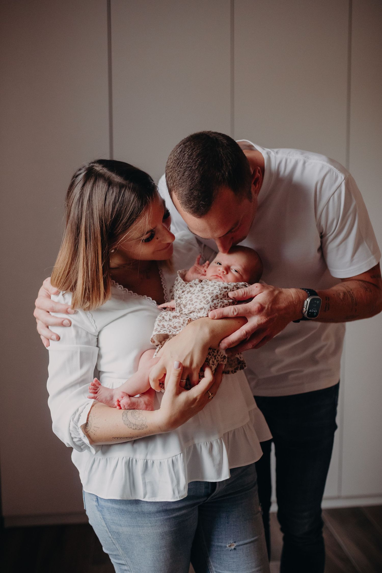 Parents in white tops share an intimate moment with their newborn baby wearing a floral outfit.