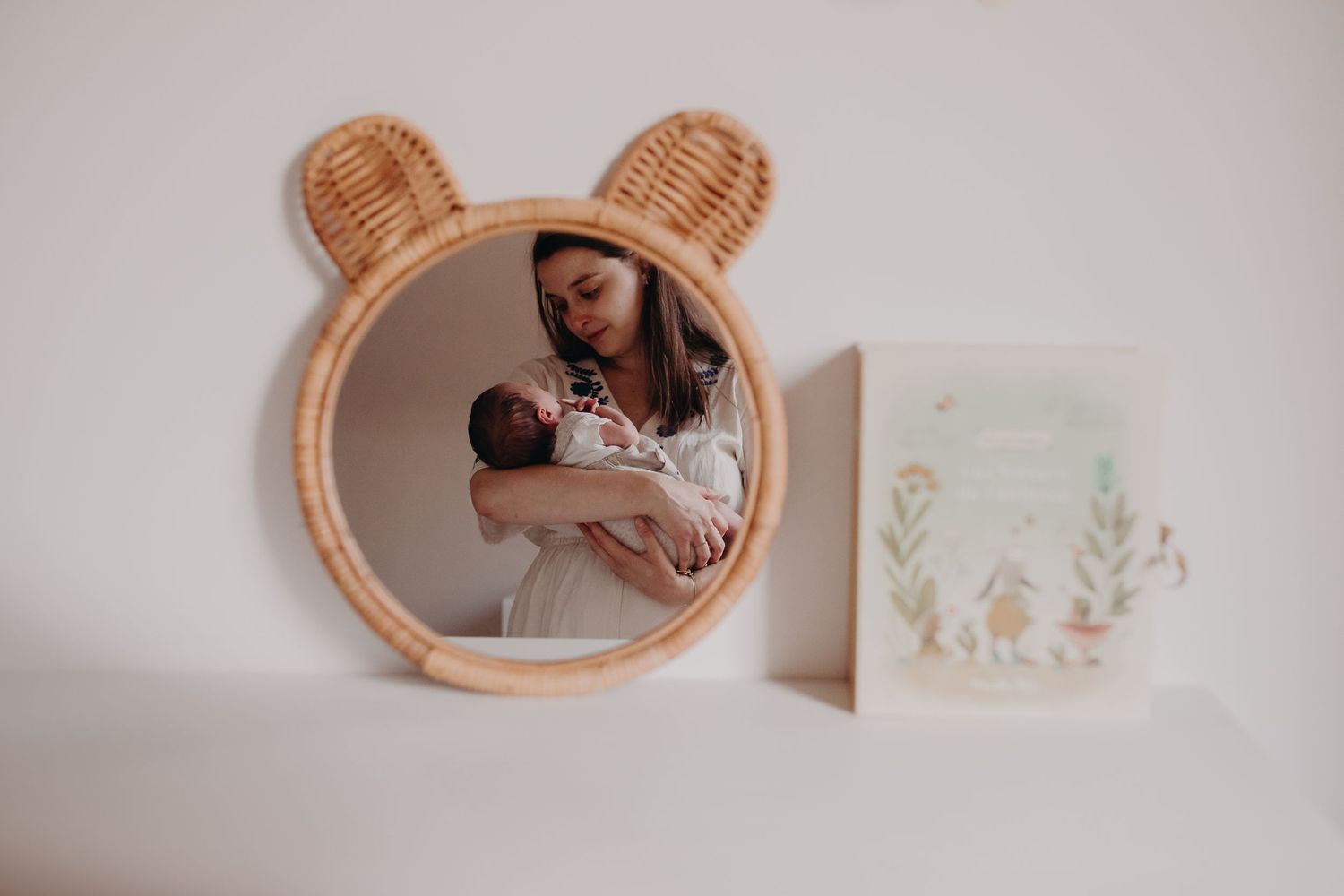 A reflection in a rattan bear-shaped mirror shows someone cradling a baby next to soft wall decor.