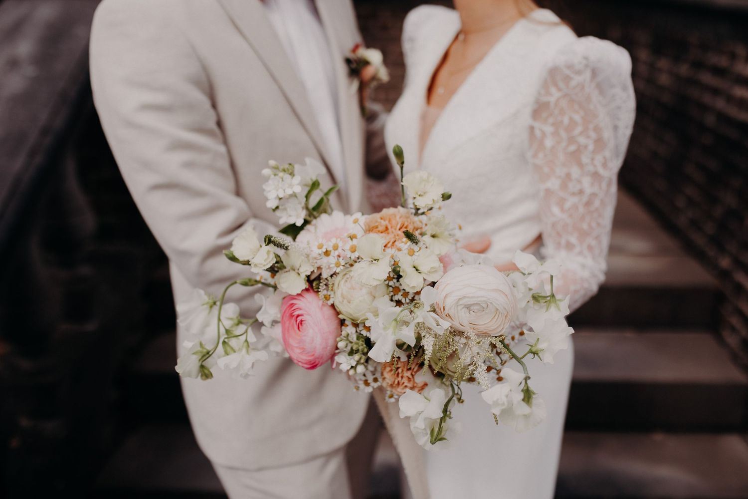 Close-up of a delicate pink and white wedding bouquet held between a couple wearing light-colored formal attire.