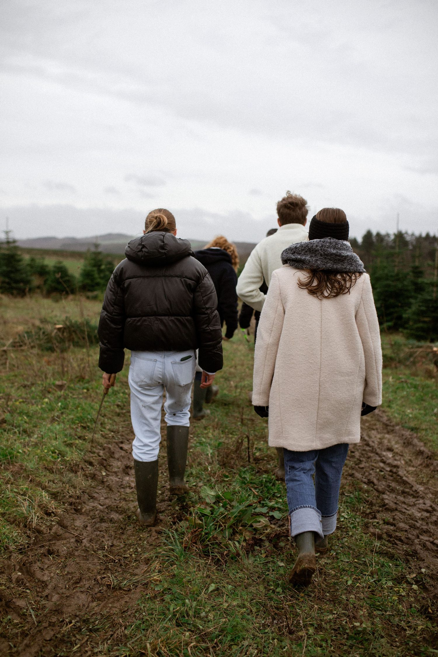 Two people walk through a muddy field in winter coats and jeans on an overcast day.